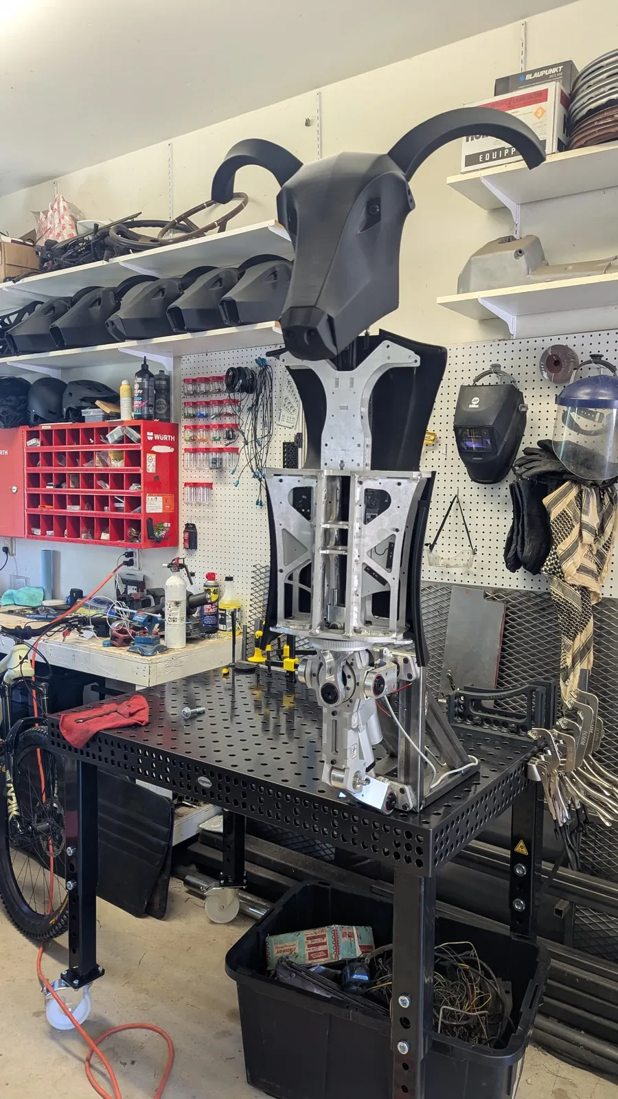 Torso incompleto do Threehalves sobre uma bancada de oficina, com estrutura metálica exposta e peças em desenvolvimento
