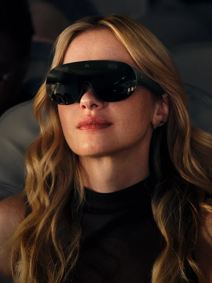 Os Meta VR Glasses concentram telas e sensores na armação; bateria e processamento ficam em um módulo externo conectado por cabo. Fonte: Meta. Mulher usa os Meta VR Glasses, que possuem visor escuro amplo e hastes mais grossas que as de óculos comuns.