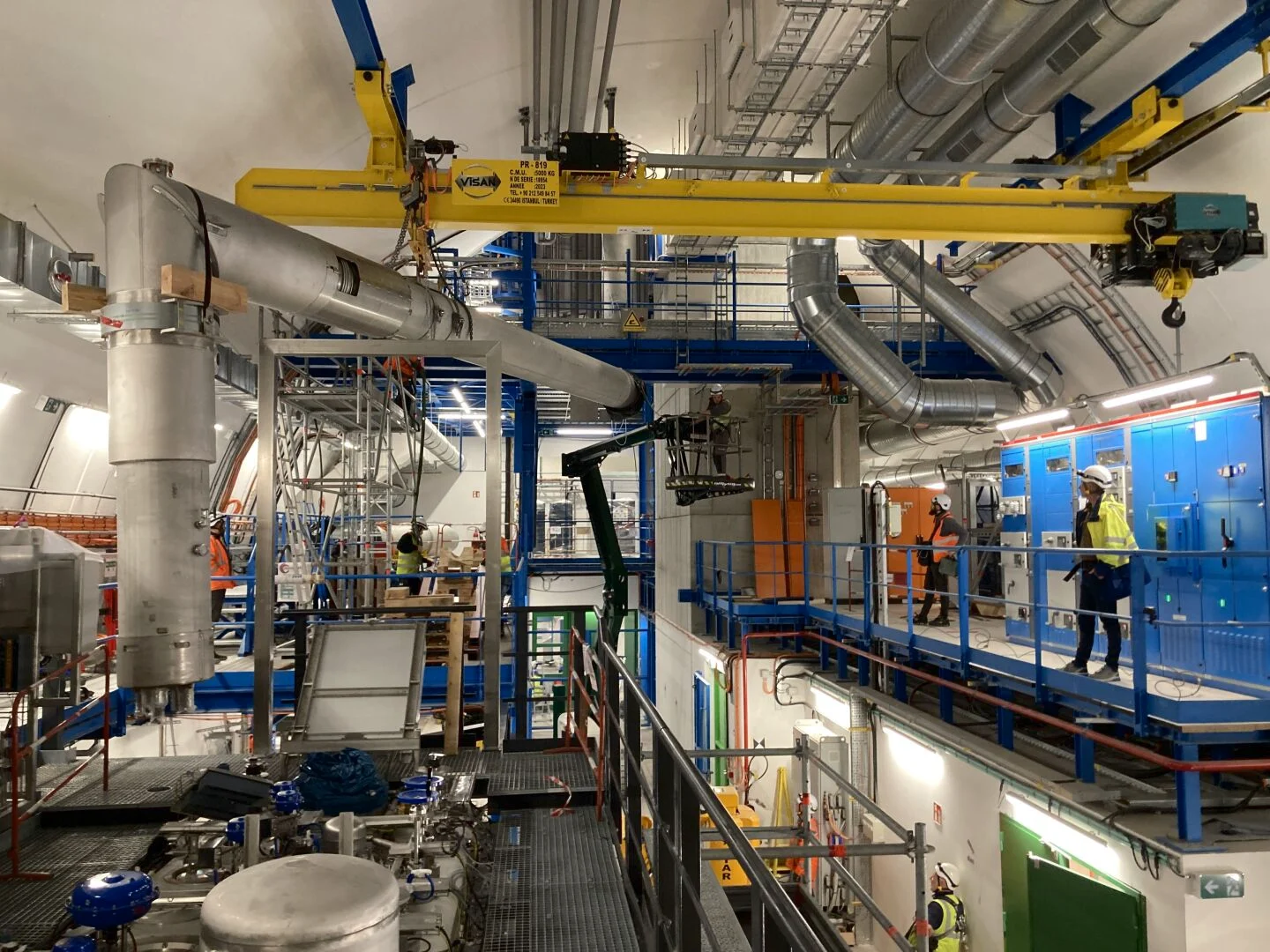 Técnicos e estruturas metálicas junto a tubos, plataformas e uma ponte rolante em uma galeria do CERN.